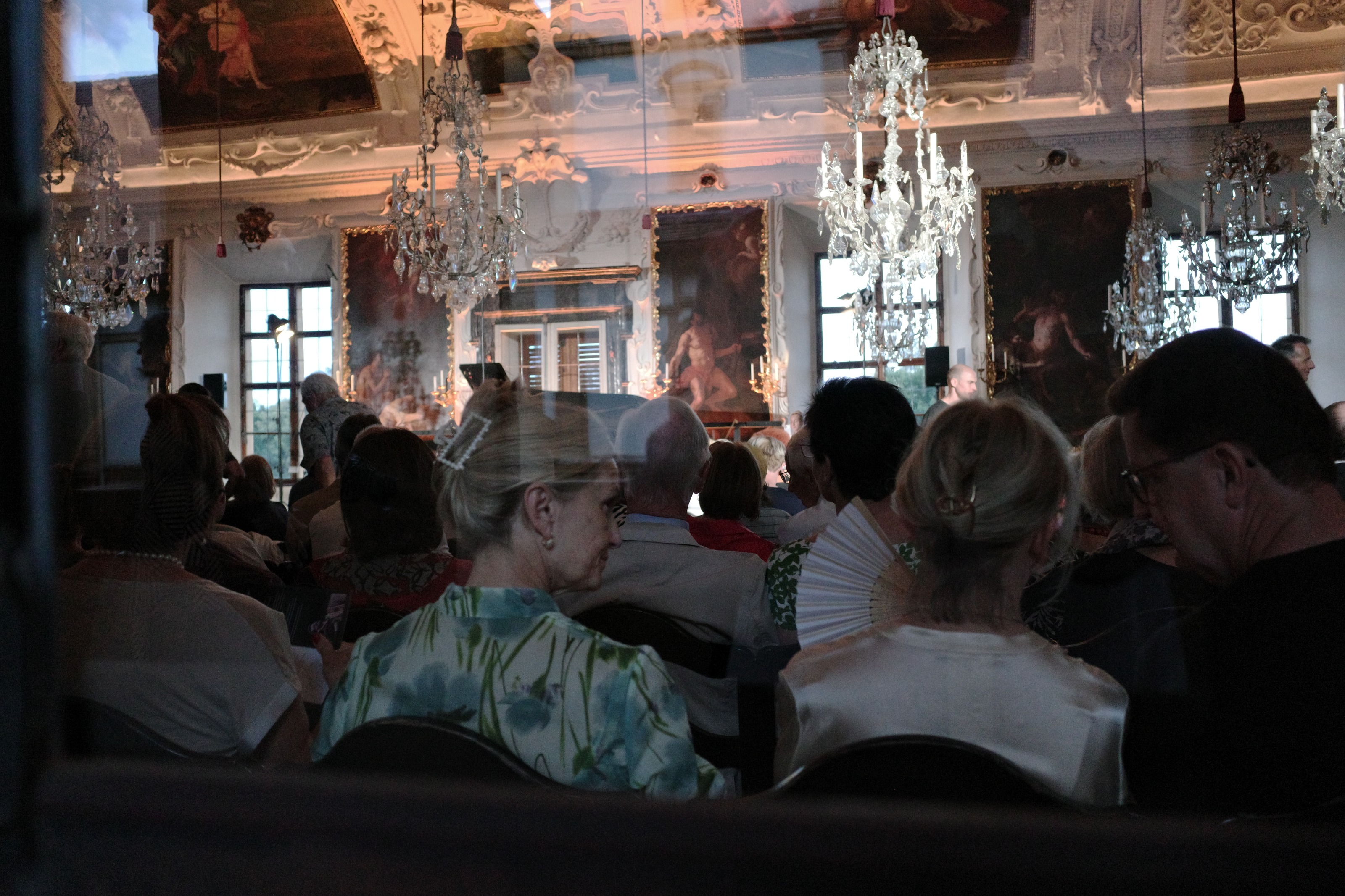 A seated audience in a frescoed hall under chandeliers, shot through a doorway