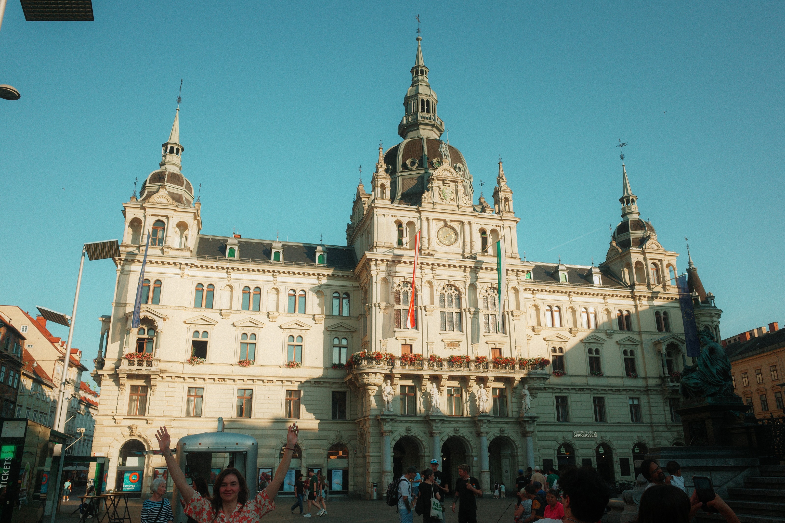 Someone throwing both arms up in front of an ornate town hall facade under a clear sky