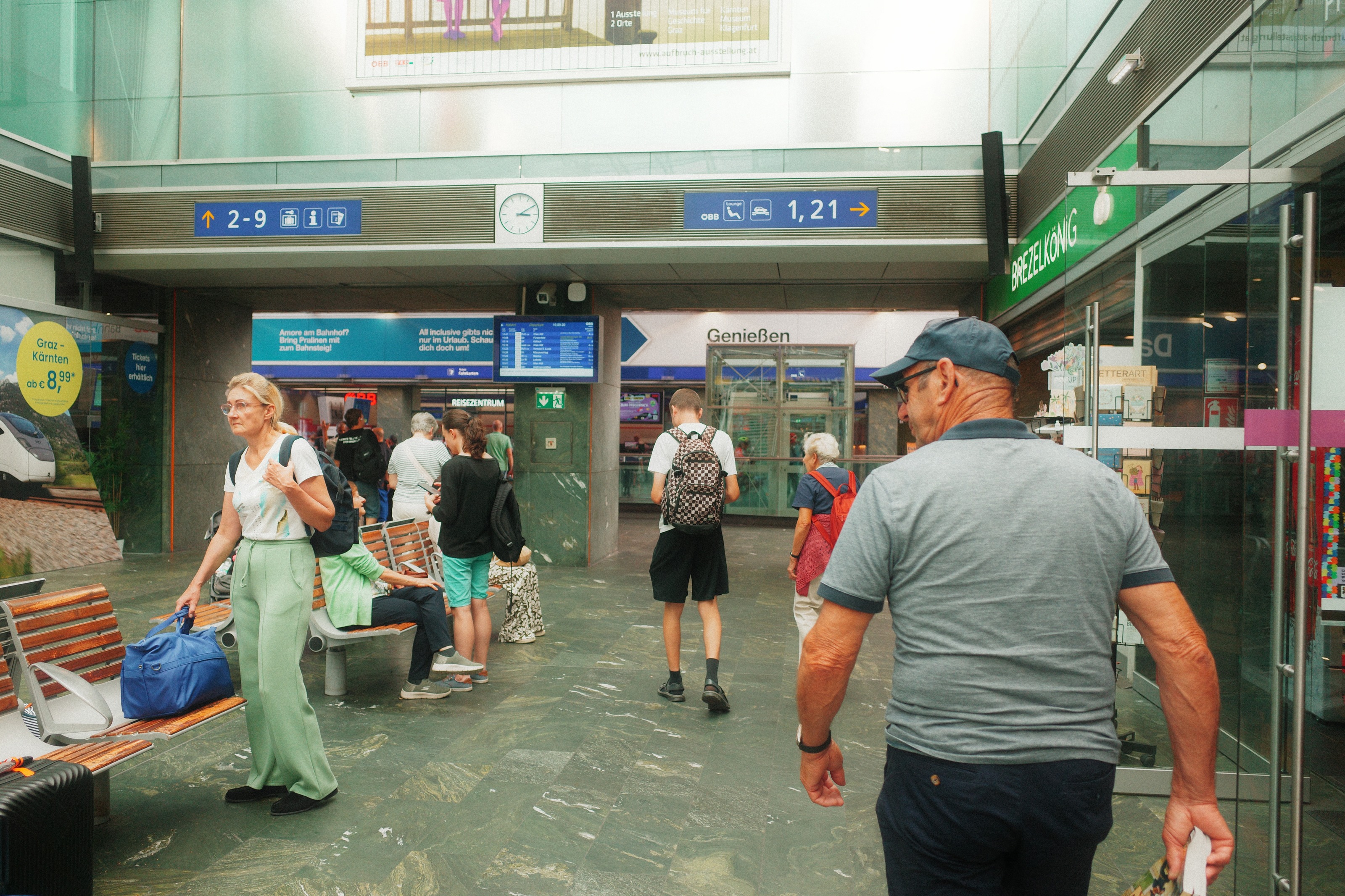 Travellers crossing a station concourse under platform signage, a man in a cap walking towards the lens